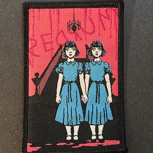 May include: A rectangular patch featuring two girls in blue dresses holding hands, set against a red background with the word "REDRUM" in red. A black border frames the design, evoking a horror theme.