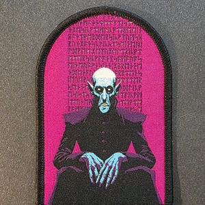 May include: Embroidered patch featuring a pale-skinned, blue-fingered figure in a black coat, set against a pink background with ancient runes. The patch has a black border and a gothic aesthetic.