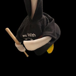 Może przedstawiać: Biała gęś w czarnym swetrze z kapturem z nadrukiem "Less With More" z przodu. Gęś trzyma drewniany kij w prawym skrzydle.