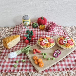 May include: A set of crocheted food items, including a pizza, a can of tomatoes, a block of cheese, a tomato plant, a red pepper, a green pepper, a baguette, and a slice of salami. The items are arranged on a red and white checked tablecloth.