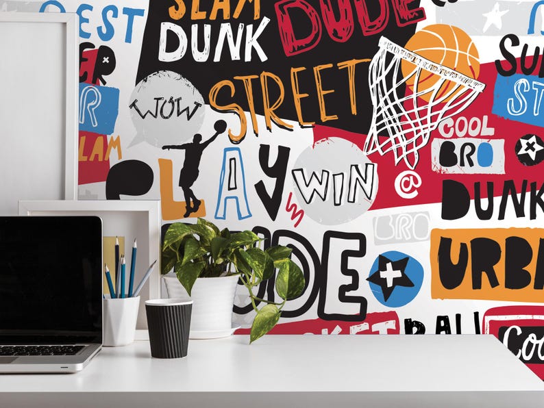 a white desk with a laptop, a cup, and a potted plant. The background behind the desk is covered in a colorful, graffiti-style wall with various words and phrases related to basketball, such as "SLAM", "DUNK", "STREET", "LAW", "WIN", "@", "DUNK", "URBAN", "COOL", "BRO", and "BALLS".