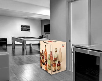 May include: A black and white image of a basement with a pool table, a sofa, and a small refrigerator. The refrigerator is decorated with images of various bottles. A framed picture hangs on the wall.