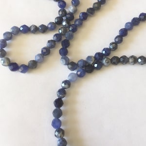 May include: A strand of blue and white gemstone beads. The beads are faceted and have a slightly irregular shape. The beads are strung on a clear thread.