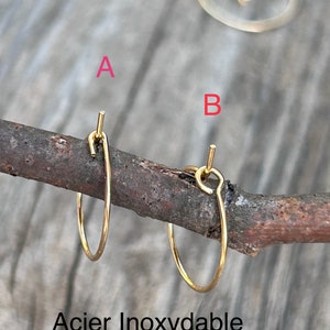 Puede incluir: Dos pendientes de aro dorados hechos de acero inoxidable. Los pendientes se muestran en una rama de madera. El texto "Acier Inoxydable" es visible debajo de los pendientes.