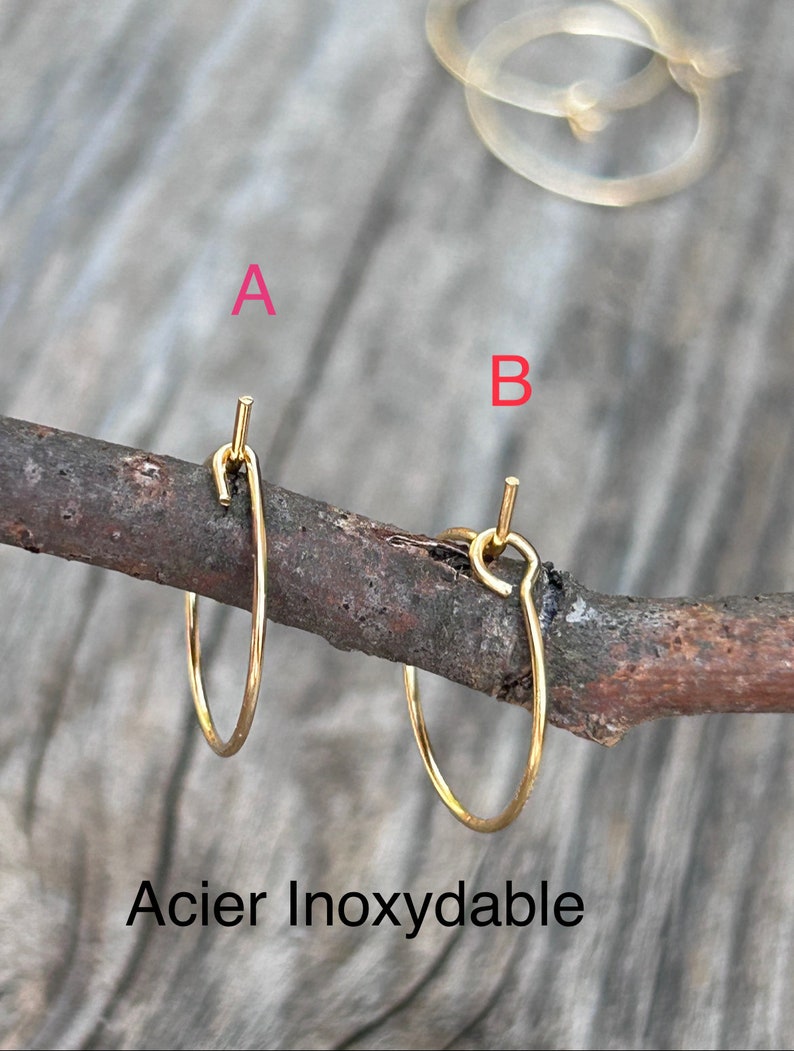 Puede incluir: Dos pendientes de aro dorados hechos de acero inoxidable. Los pendientes se muestran en una rama de madera. El texto "Acier Inoxydable" es visible debajo de los pendientes.