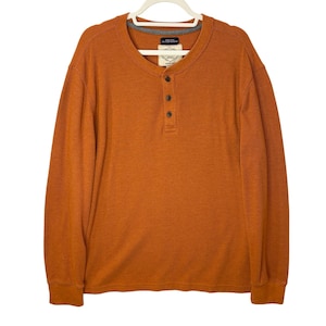 May include: An orange long-sleeve Henley shirt with a waffle knit texture. The shirt has a three-button placket and a gray trim around the neckline. The shirt is hanging on a white hanger.