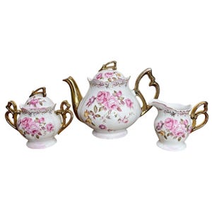 Puede incluir: Un juego de té de porcelana blanca que consta de una tetera con tapa, un azucarero con tapa y una jarra de leche. Cada pieza presenta un patrón floral de rosas rosadas, bordes festoneados dorados y asas de tono dorado.