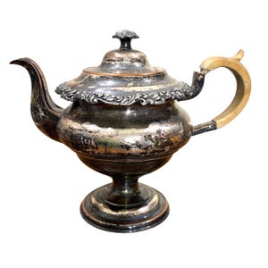 May include: Antique silver-plated teapot with a decorative floral rim and a wooden handle. The teapot has a pedestal base and a lid with a small knob. The spout curves gracefully, and the silver finish shows signs of age.