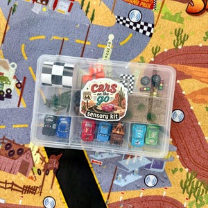 May include: A clear plastic container filled with toy cars, miniature accessories, and a checkered flag. The container is labeled "Cars on the Go Sensory Kit" and sits on a colorful play mat with a road and desert landscape.