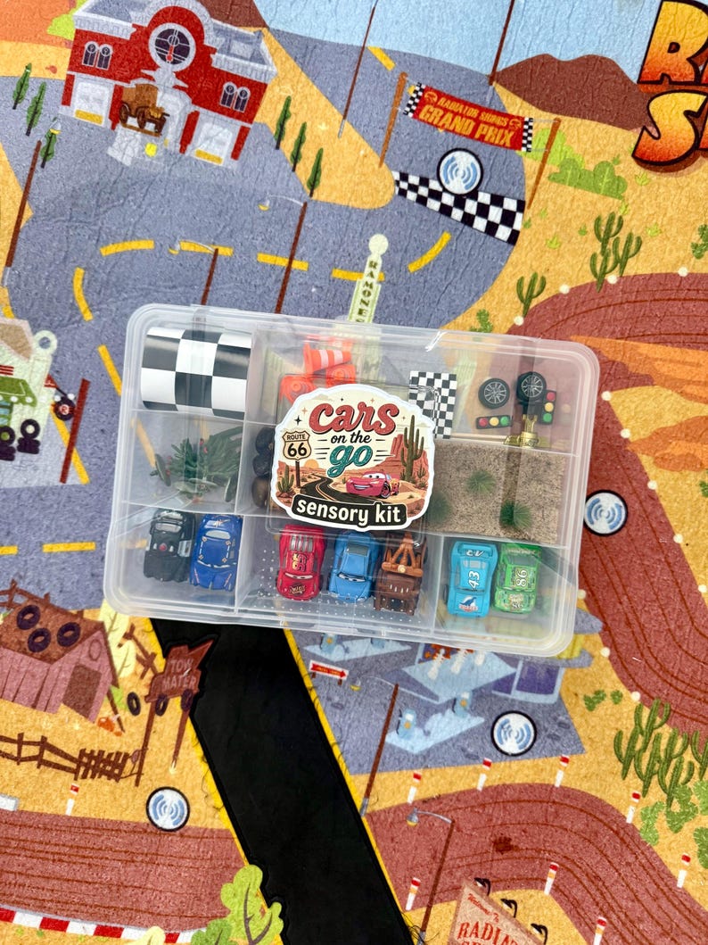 May include: A clear plastic container filled with toy cars, miniature accessories, and a checkered flag. The container is labeled "Cars on the Go Sensory Kit" and sits on a colorful play mat with a road and desert landscape.