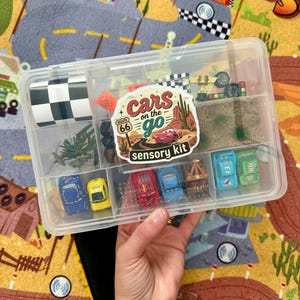 May include: A clear plastic container filled with miniature toy cars and accessories. The container is labeled "Cars on the Go" with a Route 66 theme. Includes a checkered flag, miniature trees, and other racing-themed items.