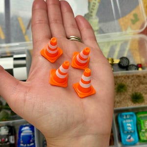 May include: Five miniature orange and white traffic cones. The cones are small, with a square base and a conical shape, and are designed with white stripes. The cones are in a clear plastic container with toy cars.