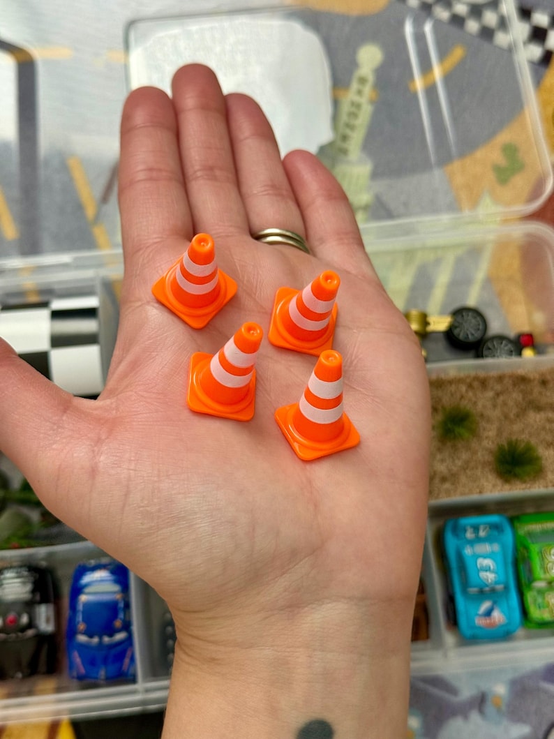 May include: Five miniature orange and white traffic cones. The cones are small, with a square base and a conical shape, and are designed with white stripes. The cones are in a clear plastic container with toy cars.