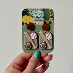 Könnte beinhalten: Ein Paar Hängeohrringe mit einem dunkelbraunen Holzkreis oben und einer grauen, bogenförmigen Basis. Die Basis zeigt einen beigen Baseballschläger, einen weißen Baseball mit roten Nähten und einen braunen Baseballhandschuh. Die Karte trägt die Aufschrift Mandy's Craft Creation.