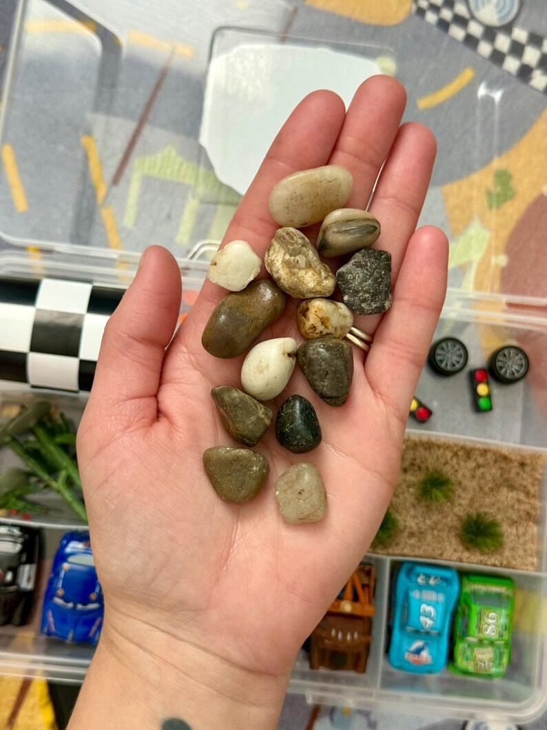 May include: A hand holding a collection of smooth, colorful stones. The stones vary in size and color, ranging from light beige to dark brown and green. The stones are in a clear plastic container with toy cars and accessories.