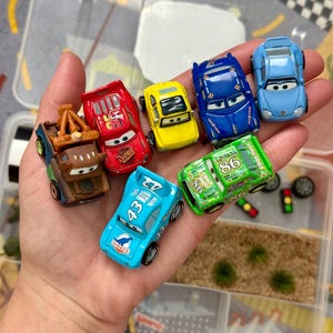 May include: A collection of toy cars in various colors, including red, yellow, blue, and green. The cars feature different designs and numbers, with some bearing the "Rust-eze" and "Dinoco" logos. The cars are small, likely made of die-cast metal or plastic.