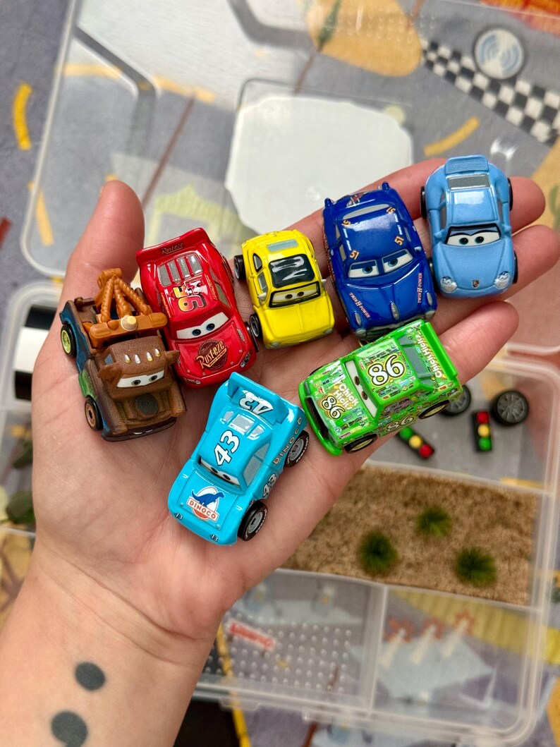 May include: A collection of toy cars in various colors, including red, yellow, blue, and green. The cars feature different designs and numbers, with some bearing the "Rust-eze" and "Dinoco" logos. The cars are small, likely made of die-cast metal or plastic.