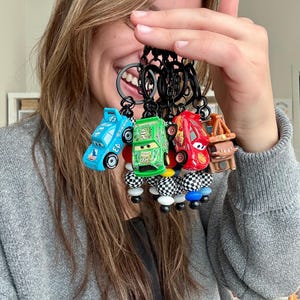 Puede incluir: Una colección de cinco llaveros de coches de juguete en varios colores, incluyendo azul, verde y rojo. Cada llavero presenta un diseño de coche diferente con anillos negros y cuentas decorativas. Los llaveros se sostienen frente a una persona.