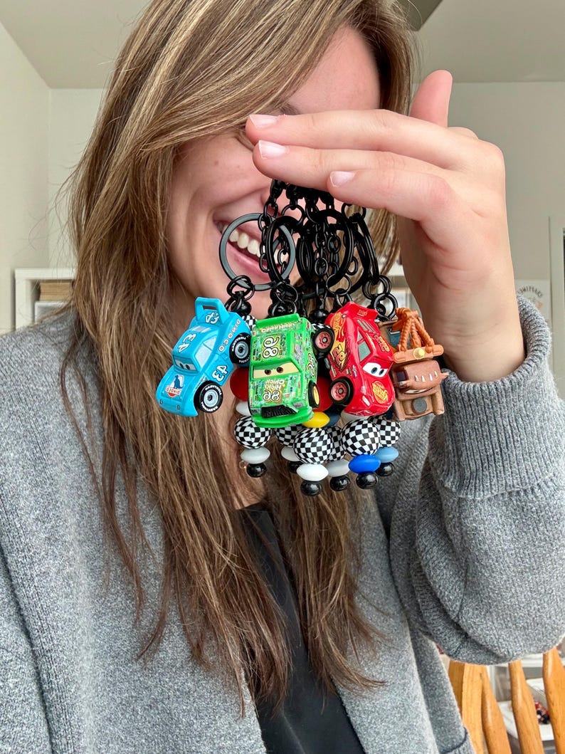 May include: A collection of five toy car keychains in various colors, including blue, green, and red. Each keychain features a different car design with black key rings and decorative beads. The keychains are held up in front of a person.