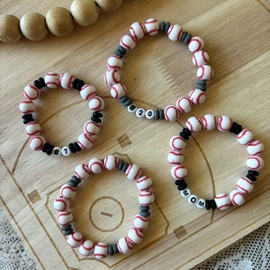 May include: Four baseball themed bracelets with gray and white beads and black lettering. The bracelets spell out "#1", "#20", "MOM", and are on a wooden surface.