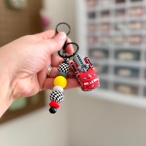 May include: A red toy fire truck keychain with a black keyring and a beaded accent. The beads are yellow, white, black and red. The fire truck has a ladder and is a cartoon style. The keychain is held by a person.
