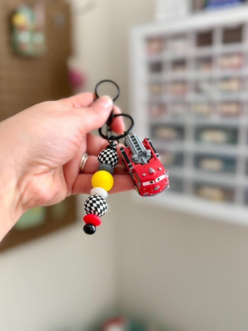 May include: A red toy fire truck keychain with a black keyring and a beaded accent. The beads are yellow, white, black and red. The fire truck has a ladder and is a cartoon style. The keychain is held by a person.