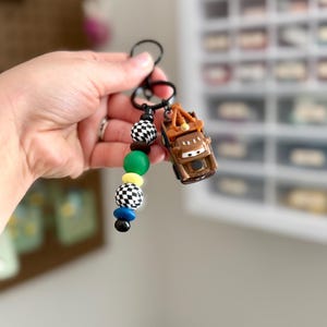 May include: A black keychain with a brown toy car and colorful beads. The beads are black and white checkered, green, yellow, blue, and black. The toy car is a brown tow truck.
