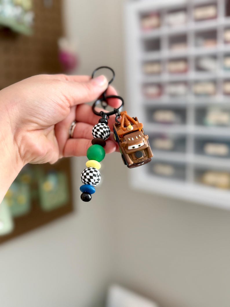 May include: A black keychain with a brown toy car and colorful beads. The beads are black and white checkered, green, yellow, blue, and black. The toy car is a brown tow truck.