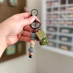 May include: A keychain with a black ring and a beaded strand in black, white, tan, and mint green. A small, olive green toy jeep with a white star is attached to the keychain.