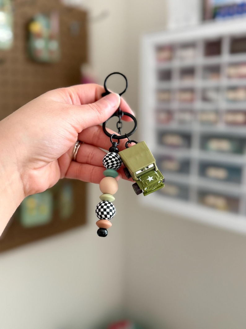 May include: A keychain with a black ring and a beaded strand in black, white, tan, and mint green. A small, olive green toy jeep with a white star is attached to the keychain.