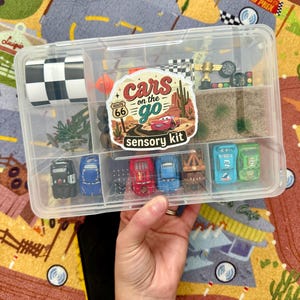 May include: A clear plastic organizer filled with toy cars and accessories. The organizer includes a sticker that says "Cars on the Go Sensory Kit". The kit contains toy cars, a checkered flag, and miniature race track elements.