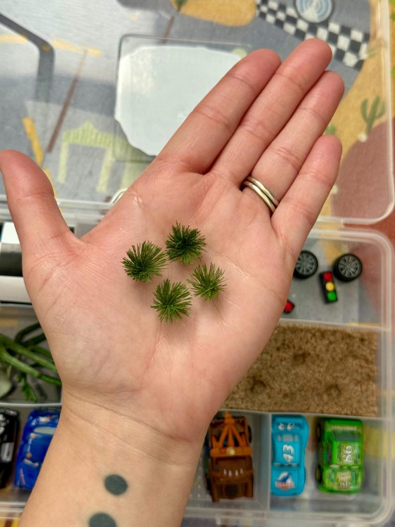 May include: A hand holding five small, green, spiky miniature trees. The hand is in a clear plastic container with toy cars, wheels, and other miniature accessories. The container has a road-themed background.