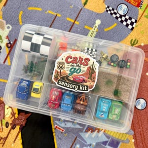 May include: A clear plastic container filled with toy cars, miniature flags, and other accessories. The container has a label that reads "Cars on the Go Sensory Kit." The background is a colorful play mat with a road and race track design.
