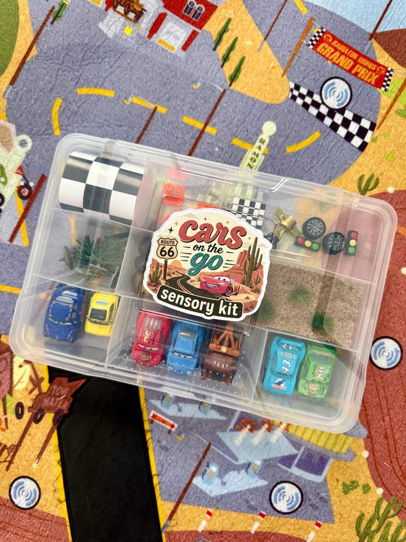 May include: A clear plastic container filled with toy cars, miniature flags, and other accessories. The container has a label that reads "Cars on the Go Sensory Kit." The background is a colorful play mat with a road and race track design.