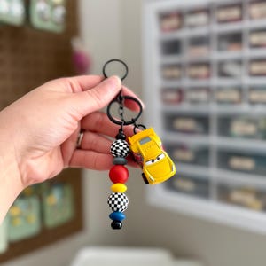 May include: A black keychain with a yellow toy car charm and colorful beads. The beads include black and white checkered, red, yellow, gray, and blue beads. The toy car is yellow with black accents.