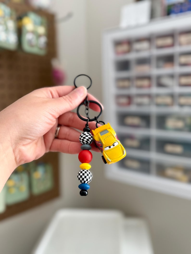 May include: A black keychain with a yellow toy car charm and colorful beads. The beads include black and white checkered, red, yellow, gray, and blue beads. The toy car is yellow with black accents.
