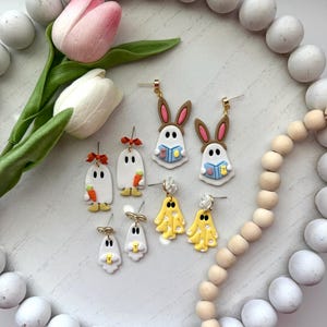May include: A collection of handmade earrings featuring whimsical designs. The earrings include ghost, bunny, and yellow ghost shapes, with details like carrots, books, and bows. The earrings are displayed on a white surface, surrounded by white and wooden beads, and a pink and white tulip.
