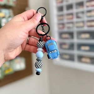 May include: A blue toy car keychain with a black keyring and a beaded accent. The accent features black and white checkered beads, light blue beads, and black beads. The keychain is held by a hand.