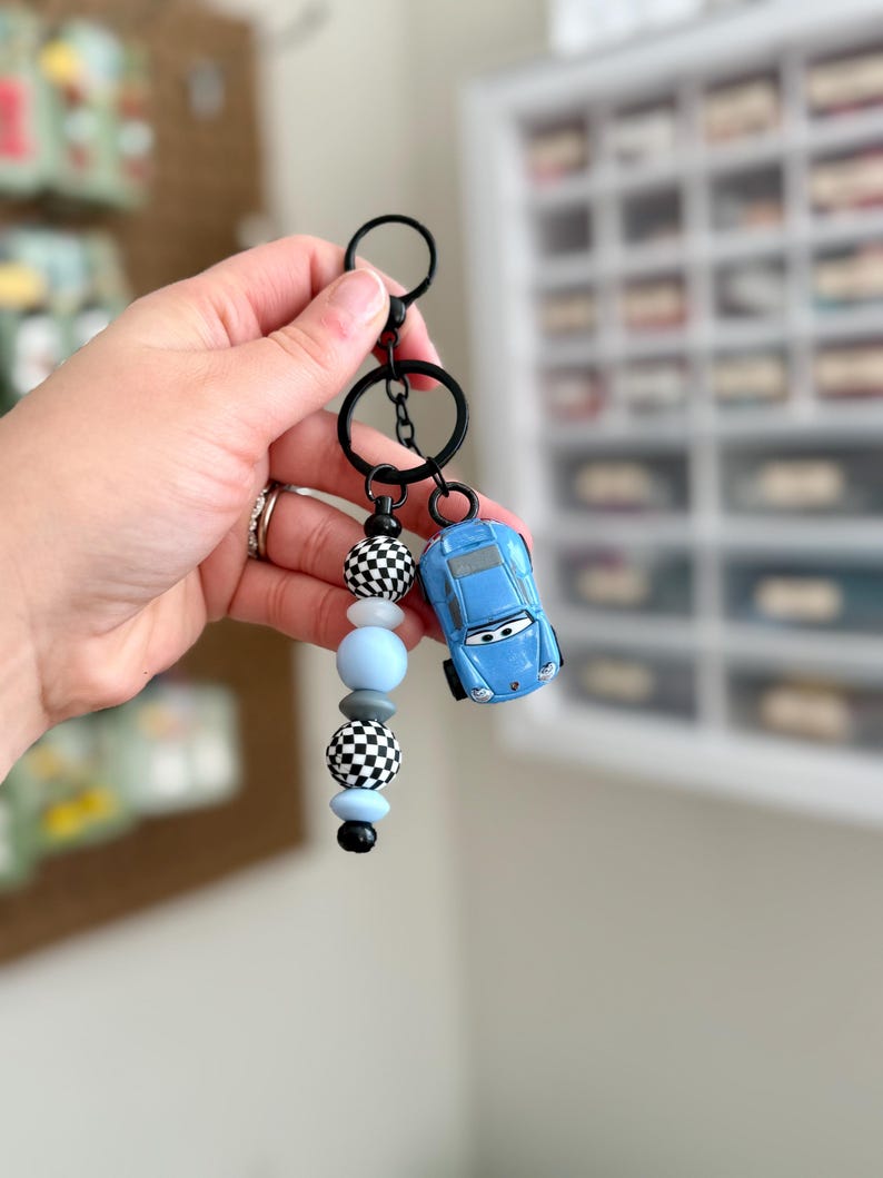 May include: A blue toy car keychain with a black keyring and a beaded accent. The accent features black and white checkered beads, light blue beads, and black beads. The keychain is held by a hand.
