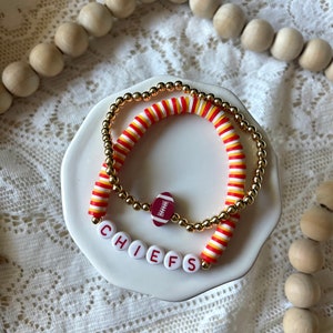Könnte beinhalten: Zwei Armbänder mit einem Football-Anhänger und dem Wort "CHIEFS" aus weißen Buchstabenperlen. Ein Armband hat goldene Perlen, das andere rot, gelb und weiß gestreifte Perlen. Die Armbänder werden auf einem weißen Teller präsentiert.