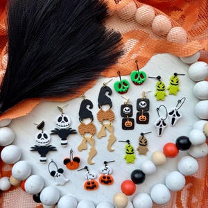 Könnte beinhalten: Eine Sammlung von Halloween-Ohrringen in verschiedenen Designs, darunter Geister, Kürbisse und Hexenhüte. Die Ohrringe sind in Schwarz, Weiß, Orange und Grün gehalten. Sie werden auf einer weißen Oberfläche mit orangefarbenem Stoff und Holzperlen präsentiert.