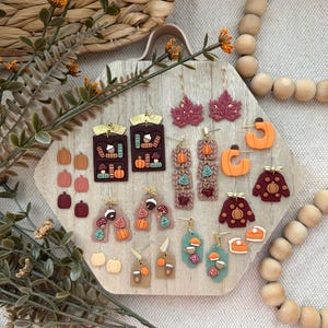 May include: A selection of autumn-themed earrings in diverse shapes and colours. The earrings feature pumpkins, leaves, books, and sweaters. The colour scheme includes orange, brown, and burgundy. Some earrings are on gold-coloured hoops or posts.