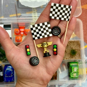 May include: Miniature racing-themed accessories, including checkered flags, a gold trophy, traffic lights, and car wheels. The items are in a clear plastic container, suggesting a craft or hobby kit. The flags are black and white.