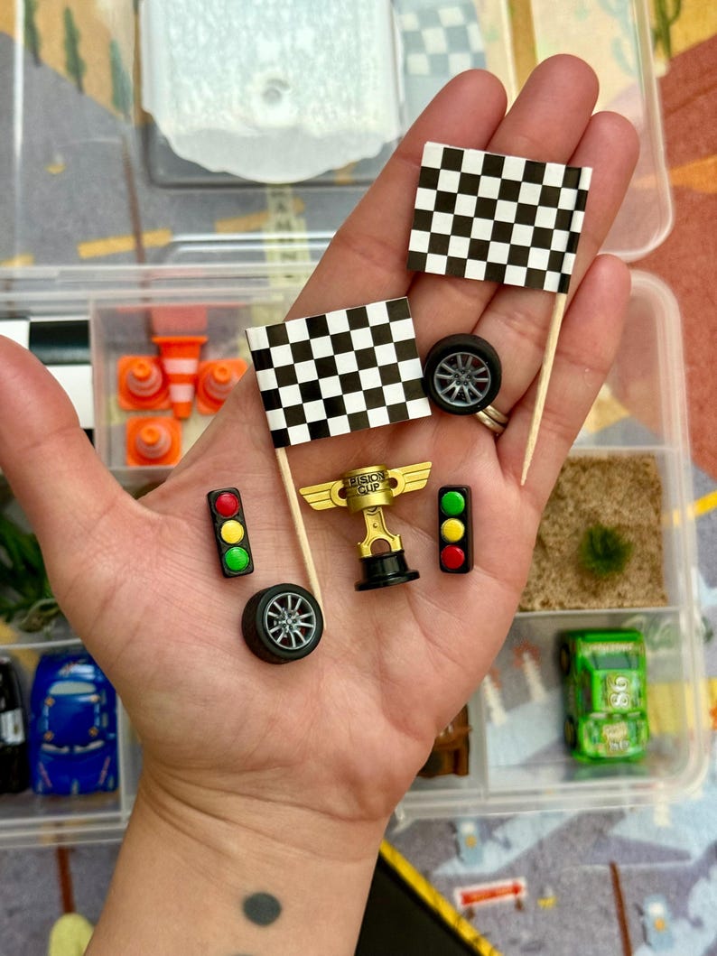 May include: Miniature racing-themed accessories, including checkered flags, a gold trophy, traffic lights, and car wheels. The items are in a clear plastic container, suggesting a craft or hobby kit. The flags are black and white.
