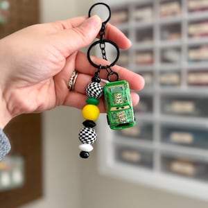 May include: A green toy car keychain with the number 86 on the side, attached to a black keyring. The keychain features a strand of black, white, yellow, and checkered beads. The car has the text "Chick Hicks" on the side.