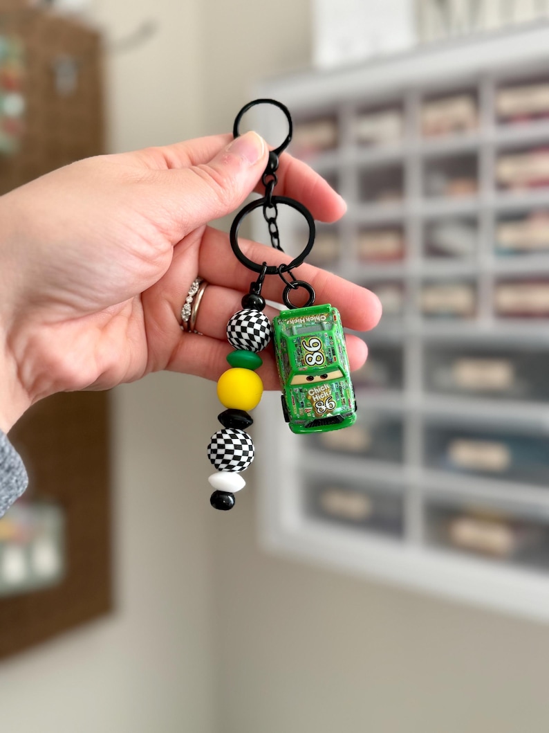 May include: A green toy car keychain with the number 86 on the side, attached to a black keyring. The keychain features a strand of black, white, yellow, and checkered beads. The car has the text "Chick Hicks" on the side.