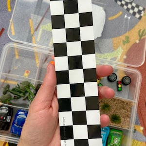 May include: A black and white checkered flag sticker is held in front of a clear plastic container with toy cars and accessories. The sticker has the text "&copy;2024 Becker Silver Design Studio". The container has miniature cars, trees, and other racing-themed items.