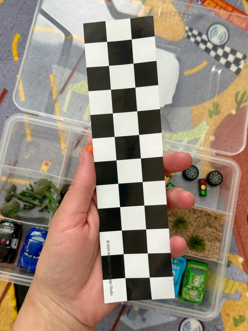 May include: A black and white checkered flag sticker is held in front of a clear plastic container with toy cars and accessories. The sticker has the text "&copy;2024 Becker Silver Design Studio". The container has miniature cars, trees, and other racing-themed items.