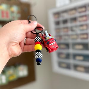 May include: A red toy car keychain with a black keyring. The keychain features a series of colorful beads, including black and white checkered, yellow, black, and blue beads. The toy car has a ladder on top.