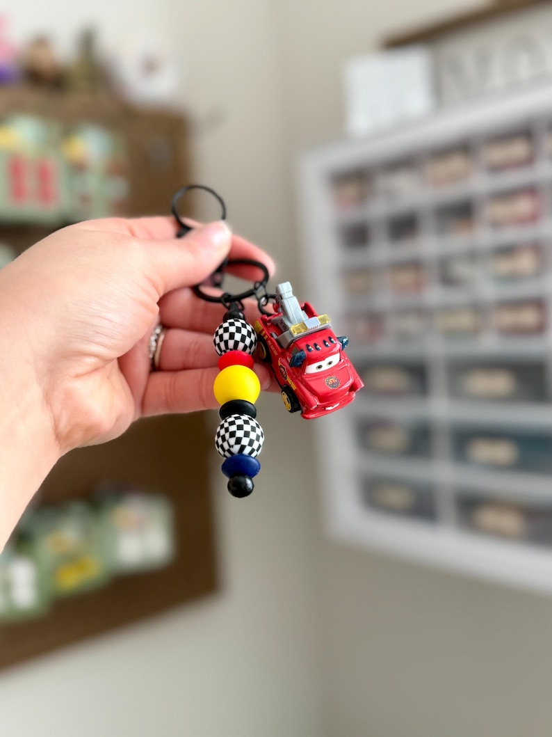 May include: A red toy car keychain with a black keyring. The keychain features a series of colorful beads, including black and white checkered, yellow, black, and blue beads. The toy car has a ladder on top.